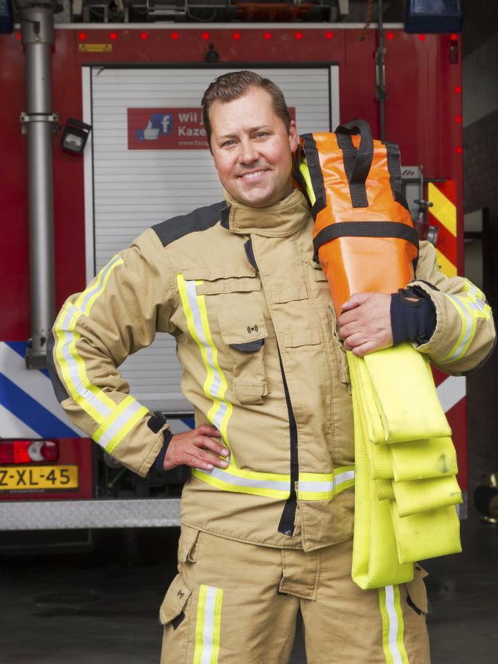Brandweerman Christiaan poseert voor brandweerwagen met blusslang op zijn schouder